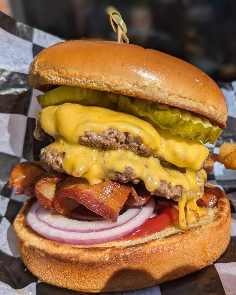Close-up of a double cheeseburger with bacon, red onion, pickles, and ketchup on a checkered wrapper.