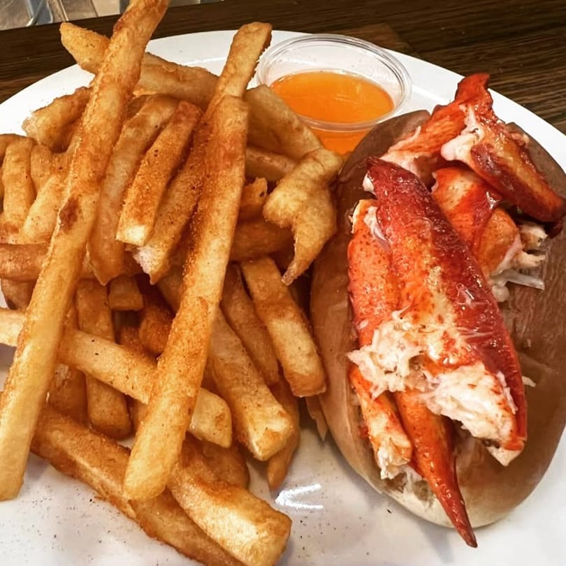 A lobster roll with large pieces of lobster meat, served with a side of french fries and dipping sauce.